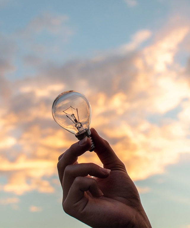 A hand holding a lightbulb with the sky in the background