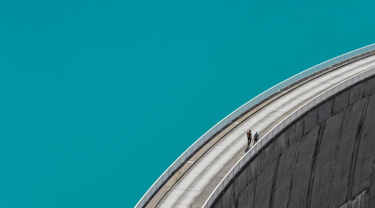 A road and sidewalk on top of a dam wall with two people walking on the sidewalk