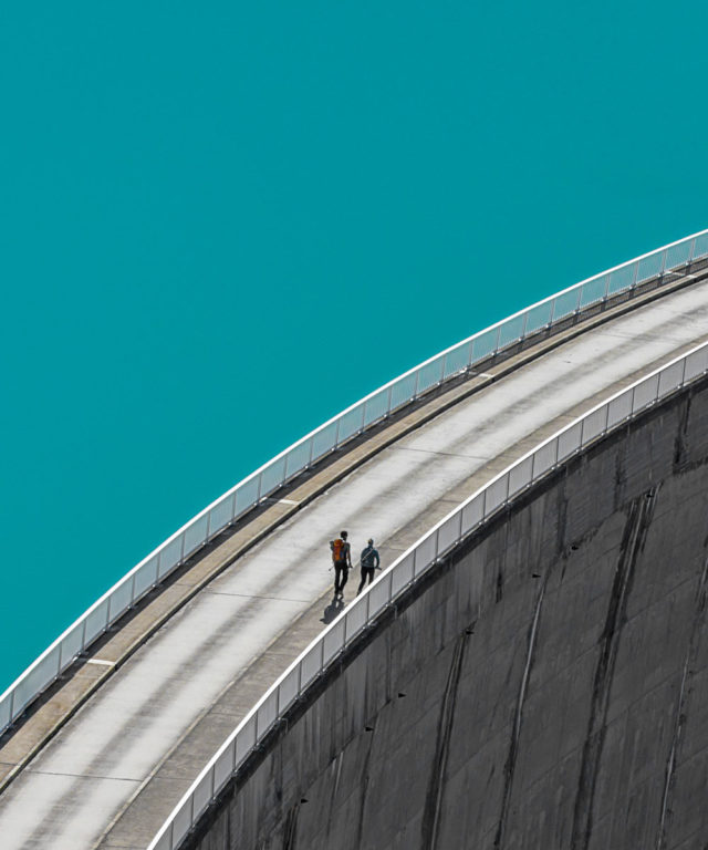 A road and sidewalk on top of a dam wall with two people walking on the sidewalk