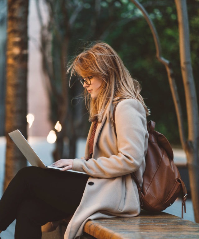 A woman sitting on a bench typing on a laptop that's open on her lap