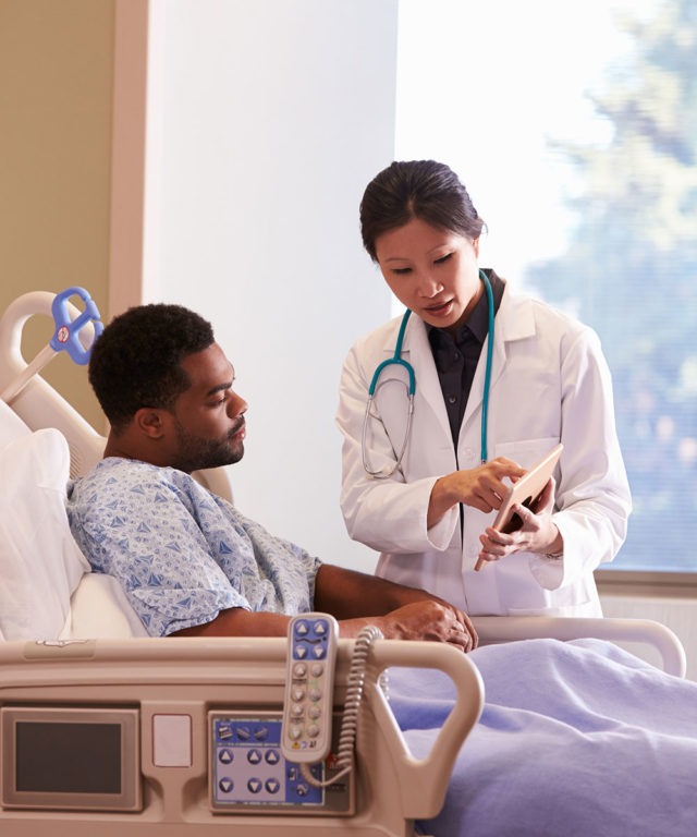 A patient in a medical bed with a doctor standing bedside, looking at a small tablet
