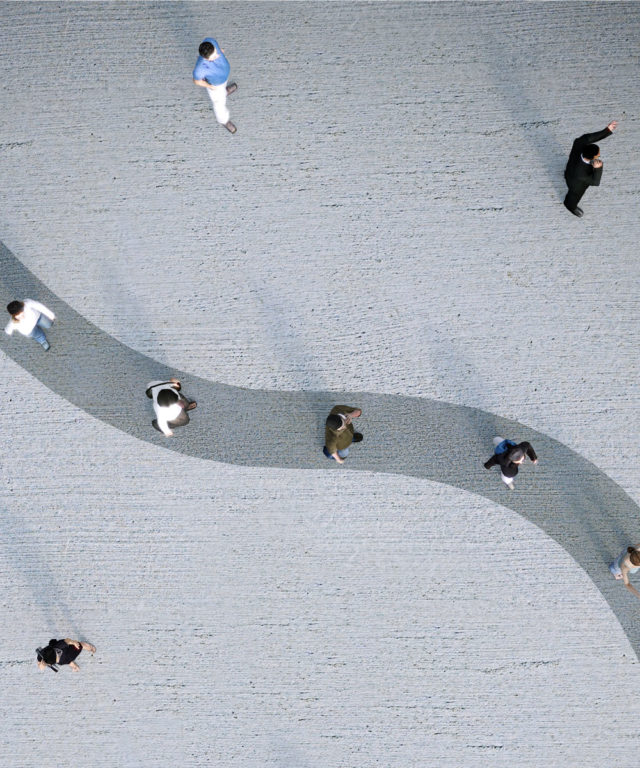 Bird's-eye view of people in a line walking along a dark grey path and other people standing around in a light grey area