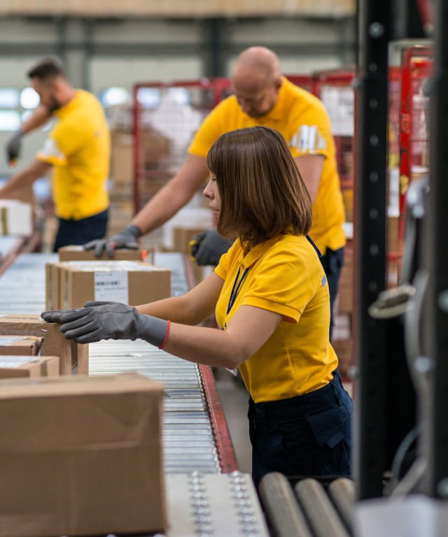 A woman touching a cardboard box on an assembly line with other workers in the background