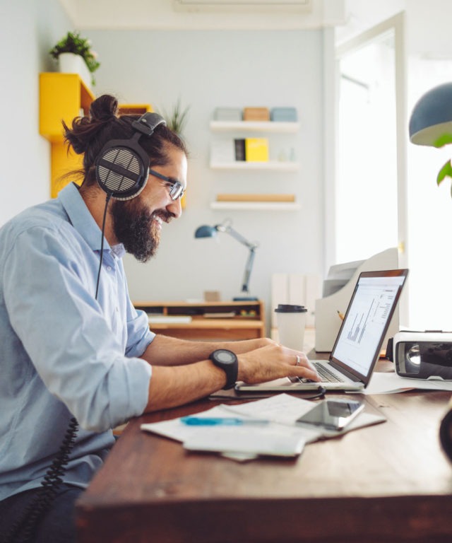 A man smiling with headphones on, sitting at a desk while working on a laptop