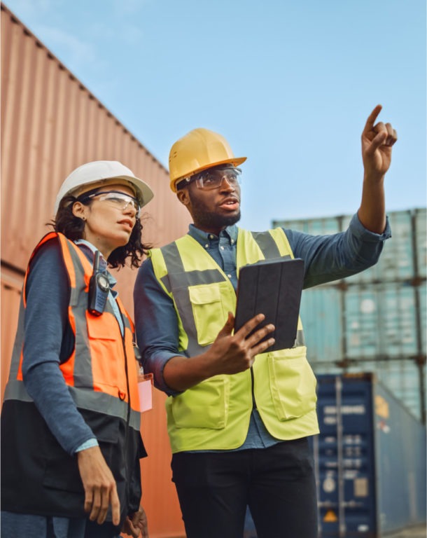 Two engineers in hard hats surrounded by cargo, on pointing while holding a tablet