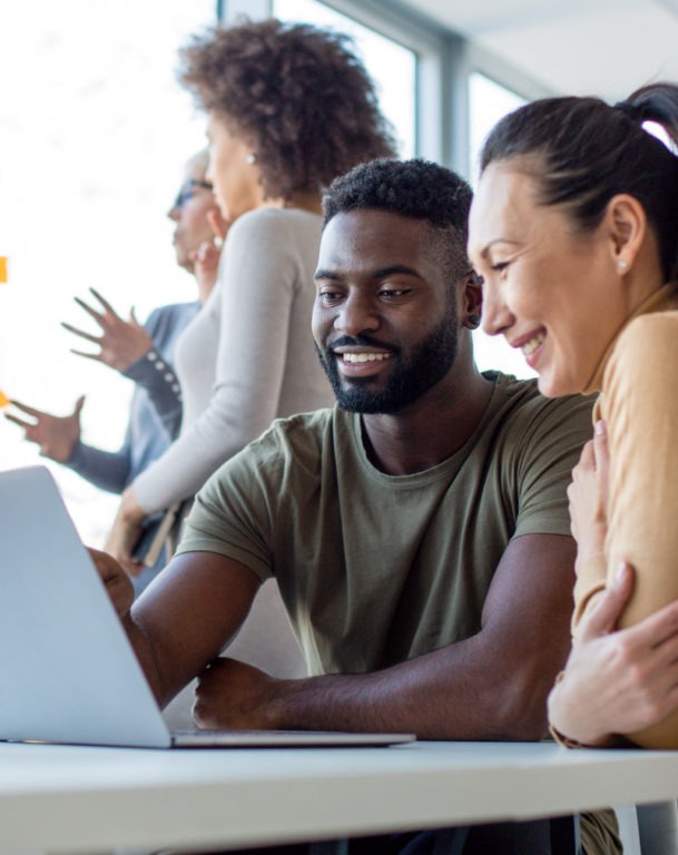 Two coworkers viewing laptop while three colleagues brainstorm in the background