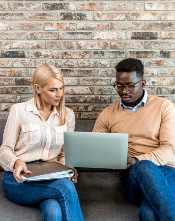 Two coworkers looking at laptop while sitting on couch against a brick wall