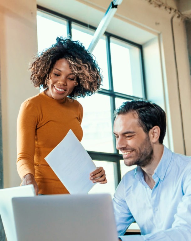 Two smiling colleagues in front of laptop, one standing while the other is seated