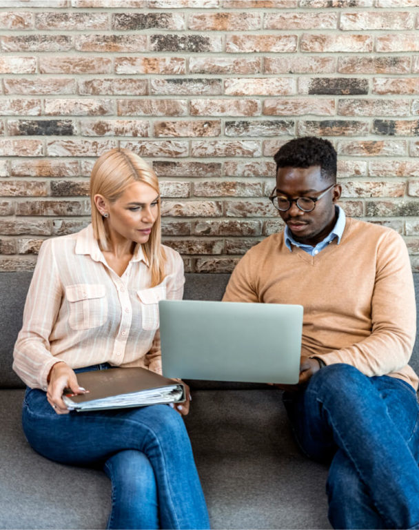 Two coworkers looking at laptop while sitting on couch against a brick wall